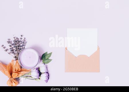 Umschlag mit leerer weißer Blattschablone neben Lavendelblüten, Duftkerze und parfümiertem Beutel auf hellem lila Hintergrund, flaches Lay. Stockfoto