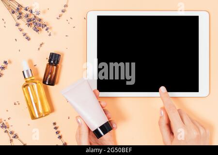 Flach legen weibliche Hände halten Cremeböhrchen und mit Tablette mit leerem Bildschirm neben Bio-Hautpflegeprodukte auf beigem Hintergrund mit Lavendel Flowe Stockfoto