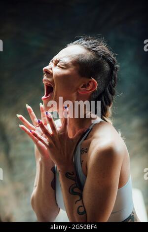 Eine junge Frau in einer Gymnastik, die vor einem Hintergrund steht. Sie schreit laut und ihre Hände sind ausgestreckt. Stockfoto