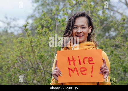 Eine junge Frau, die draußen vor Bäumen steht und ein Stück Papier mit „hire me“ darauf hält. Sie blickt auf die Kamera und lächelt. Stockfoto