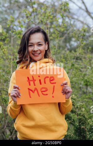 Eine junge Frau, die draußen vor Bäumen steht und ein Stück Papier mit „hire me“ darauf hält. Sie blickt auf die Kamera und lächelt. Stockfoto