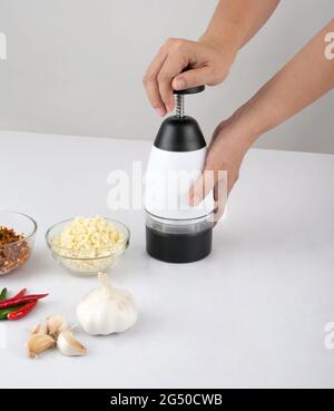 Frau hackt Knoblauch und Chili mit einem Zerkleinerer-Werkzeug auf dem weißen Tisch in der Küche Stockfoto