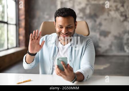 Fröhlicher inder, der per Videoanruf auf dem Smartphone spricht, auf dem Stuhl in modernen Büroflächen sitzt, Hallo in die Webcam winkt, glücklich, Freunde oder Familie auf der Ferne zu treffen Stockfoto