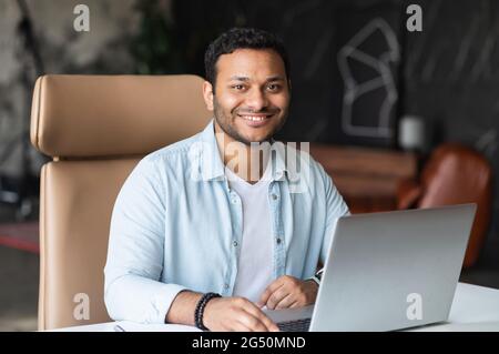 Optimistischer, selbstbewusster, dunkelhaariger indischer, hübscher Mann, der ein legeres Hemd trägt und einen Laptop für die Arbeit im Büro verwendet, sitzt ein freiberuflicher Mann aus dem Osten mit gemischter Rasse am Schreibtisch und blickt auf die Kamera und lächelt freundlich Stockfoto