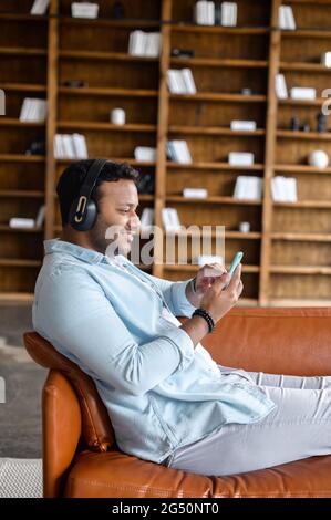 Attraktiver junger indischer Hipster, der Kopfhörer trägt, sich entspannt und auf dem bequemen Sofa liegend, hält Smartphone, hört Musik, chattet in den sozialen Medien auf dem Handy, schaut Filme Stockfoto