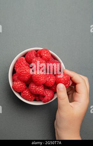 Himbeeren in einer Untertasse auf schwarzem Hintergrund. Das Kind nimmt Himbeeren vom Teller. Schiefersteinstruktur. Vertikales Foto Stockfoto