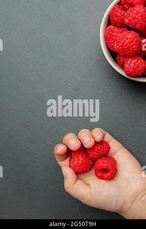 Himbeeren in einer Untertasse auf schwarzem Hintergrund. Das Kind nimmt Himbeeren vom Teller. Schiefersteinstruktur. Vertikales Foto Stockfoto