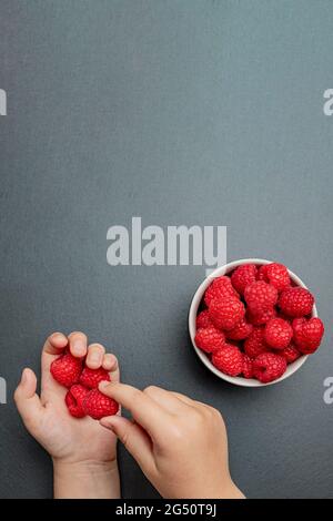 Himbeeren in einer Untertasse auf schwarzem Hintergrund. Das Kind nimmt Himbeeren vom Teller. Schiefersteinstruktur. Vertikales Foto Stockfoto