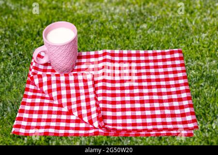 Leere Frühstückdecke. Nahaufnahme eines rosa Keramikbechers mit Milch auf rot karierter Serviette oder Tischdecke auf verschwommenem, sonnenüberflutetem, saftigem Gras. Wunderschönes BA Stockfoto