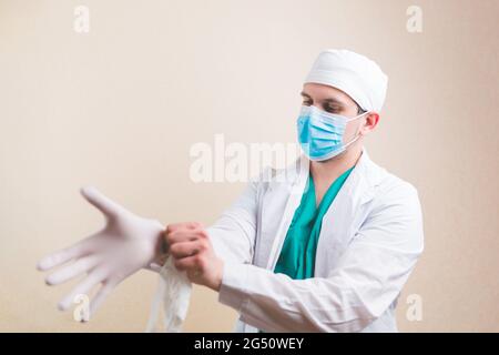 Der junge Arzt in weißem Anzug und schützender Atemmaske legt Handschuhe auf seine Hände auf gelben Hintergrund isoliert. Motiom Unschärfe. Selektiver Fokus. Stockfoto