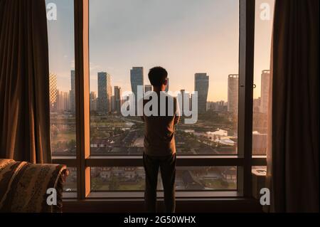 Junger asiatischer Mann, der in einem Hotel durch das Fenster schaut. Sonnenaufgang über modernen Gebäuden im Songdo Central Park in Incheon, Südkorea Stockfoto