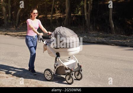 Junge Frau trägt legere Kleidung und Sonnenbrille schieben Baby Wagen Kinderwagen auf Asphalt Straße, sonnigen Tag, Wald im Schatten Hintergrund Stockfoto