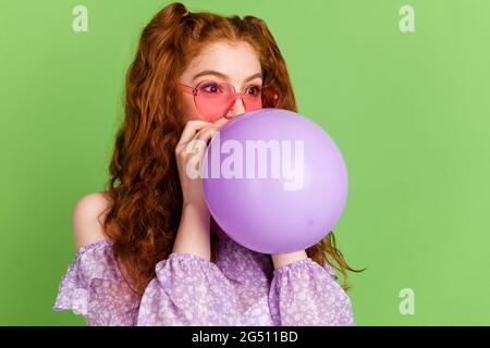 Foto von süßen roten Frisur junge Dame Schlag Ballon suchen leeren Raum tragen violette Bluse Brille isoliert auf grünem Hintergrund Stockfoto