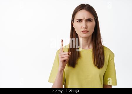 Nahaufnahme des Mädchens macht verwirrtes Gesicht, nicht bekommen, zeigt nach oben, runzelte die Stirn und schielte verwirrt, kann nicht verstehen smth, gegen Weiß stehen Stockfoto