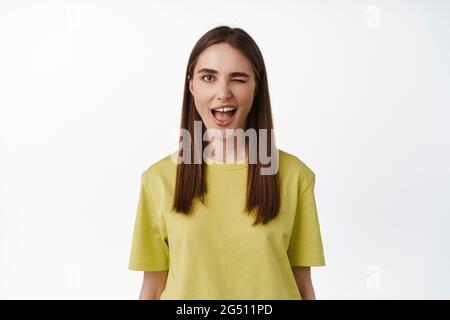 Positives Mädchen zwinkert dich an, deutet auf smeth, lächelnd und glücklich, stehend in gelbem T-Shirt vor weißem Hintergrund Stockfoto