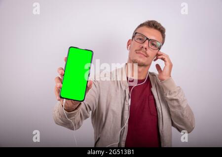 Junger attraktiver Mann hört Musik mit Kopfhörern, zeigt seinen Smartphone-Bildschirm, grünen Bildschirm, um sein Logo einzufügen, Studiofoto. Stockfoto