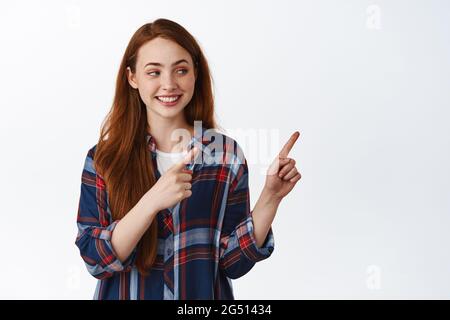 Natürliches Mädchen Teenager mit roten Haaren, zeigt und schaut nach rechts, zeigt Kopieplatz, Werbung etwas, stehend in karierten Hemd gegen weiß Stockfoto