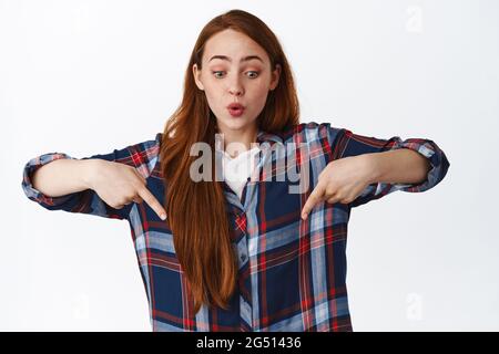 Überrascht und amüsiert Ingwermädchen, kaukasische Frau zeigte Finger nach unten und Blick auf Shopping-Werbung, stehend in karierten Hemd über weiß Stockfoto