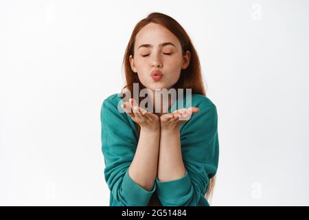 Zarte und schöne Rotschopf Frau Pucker Lippen, senden Luftkuss in offenen Händen, stehen romantisch und niedlich vor weißem Hintergrund Stockfoto