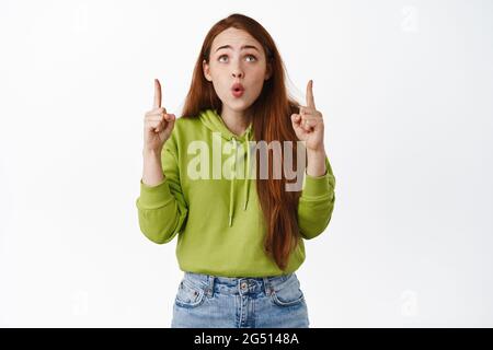 Porträt einer verblüfften Rotschopf-Frau mit langen Haaren, Seufzen fasziniert, zeigt und schaut erstaunt auf, sagen wow, beeindruckt von etwas kühlem, weißem Stockfoto