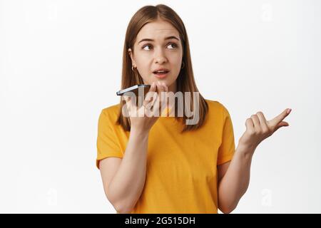 Frau zählt, während sie auf dem Freisprechtelefon ist, auf dem Mobiltelefon diktiert, denkt und benennt smeth, zeichnet Sprachnachrichten auf, steht in gelbem T-Shirt über Weiß Stockfoto