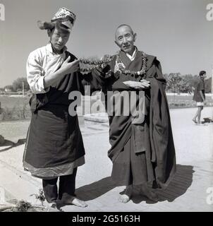 1950er Jahre, historisches Foto von J Allan Cash von einem Jugendlichen in Roben und Hut, der mit einem Mönch vor dem buddhistischen Tempel stand, mulagandhakuti vihara, Sarnath, Varanasi, indien. Der junge Mann hält Blumengirlanden oder Zöpfe, die um den Hals getragen werden und die er den Besuchern anbietet. Der Tempel wurde 1931 - an der Stelle, an der Buddha Gautama während seiner ersten Regenzeit vermittelte - von Angarika Dharmapala erbaut und wird von der Mahabodhi Society, von der er gegründet hat, unterhalten und geführt. Stockfoto