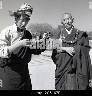 1950er Jahre, historisches Foto von J Allan Cash von einem Jugendlichen in Roben und Hut, der mit einem Mönch vor dem buddhistischen Tempel stand, mulagandhakuti vihara, Sarnath, Varanasi, indien. Der junge Mann hält Blumengirlanden oder Zöpfe, die um den Hals getragen werden und die er den Besuchern anbietet. Der Tempel wurde 1931 - an der Stelle, an der Buddha Gautama während seiner ersten Regenzeit vermittelte - von Angarika Dharmapala erbaut und wird von der Mahabodhi Society, von der er gegründet hat, unterhalten und geführt. Stockfoto