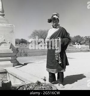 1950er Jahre, historisches Foto von J Allan Cash von einem Jugendlichen in Roben und Hut, der vor dem buddhistischen Tempel steht, mulagandhakuti vihara, Sarnath, Varanasi, indien, Hält Blumengirlanden oder Zöpfe, die um den Hals getragen werden und die er anbietet oder vielleicht an Besucher verkauft. Der Tempel wurde 1931 - an der Stelle, an der Buddha Gautama während seiner ersten Regenzeit vermittelte - von Angarika Dharmapala erbaut und wird von der Mahabodhi Society, von der er gegründet hat, unterhalten und geführt. Stockfoto