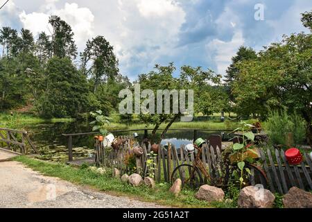 Blick auf einen kleinen See, umgeben von Bäumen, Wald im Hintergrund; alter, beschädigter Zaun mit einigen Töpfen darauf und verrosttes Fahrrad im Vordergrund Stockfoto