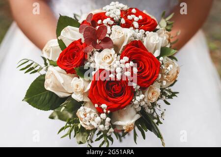 Eine Braut in einem Hochzeitskleid hält einen dekorativen Brautstrauß aus roten und weißen Rosen in ihren Händen, Nahaufnahme. Stockfoto