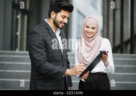 Seitenansicht des erfolgreichen arabischen Geschäftsmann Unterzeichnung Papiere auf Zwischenablage, die charmante Frau in Hijab halten. Vereinbarung über die Zusammenarbeit zwischen zwei Partnern. Stockfoto