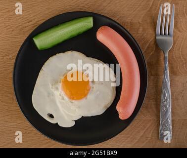 Spiegelei, Gurke und Wurst auf einem schwarzen Teller, der sich an der Stelle mit einer Gabel auf einem Holzbrett befindet Stockfoto