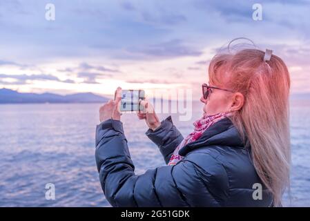 Erwachsene blonde Frau, die ihr Smartphone benutzt, um während des Sonnenuntergangs ein Foto von der Küste zu machen. Stockfoto