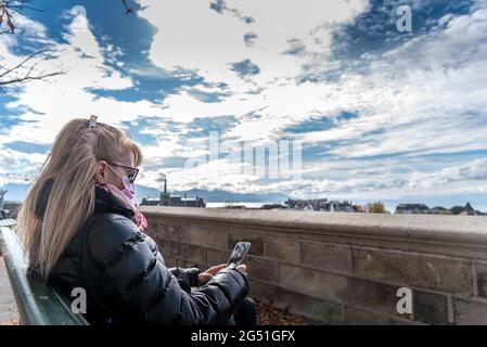 Blonde Boomerin mit Gesichtsmaske, die auf der Straße sitzt und ihr Smartphone benutzt. Stockfoto