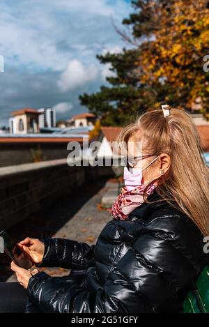 Reife blonde Frau in schwarzer Jacke, die auf der Straße sitzt und ihr Mobiltelefon benutzt. Vertikales Foto Stockfoto