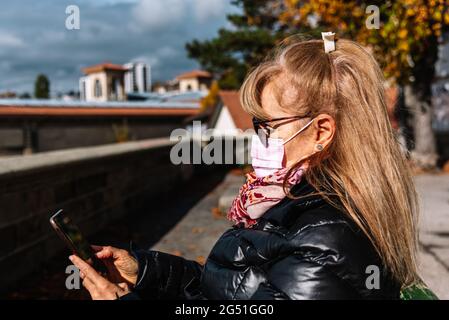 Blonde Frau mit Gesichtsmaske sitzt auf der Straße und benutzt ihr Mobiltelefon. Horizontales Foto. Stockfoto