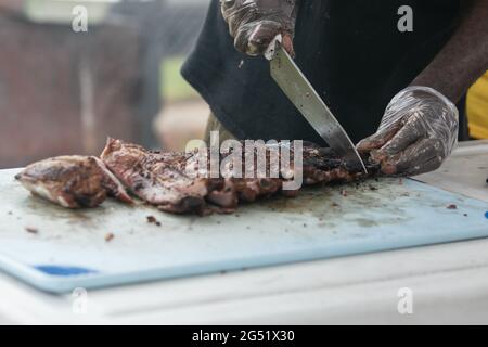 Ein Mann, der mit Handschuhen und einem großen Messer Grillrippen vom Grill auf dem Schneidebrett zerschnitt. Stockfoto