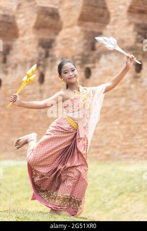 Eine langhaarige asiatische Schülerin in traditionellen thailändischen Kostümen hält silberne und goldene Blumen und führt Tanzbewegungen auf. Stockfoto