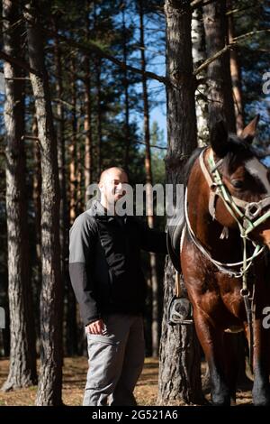 Junger Mann, der in der Nähe von Pferd steht und sich auf ein braunes Pferd vorbereitet, das über das Feld schlendert Stockfoto