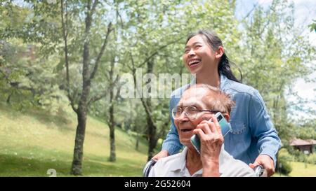 Fröhlicher, behinderter älterer Mann, der auf dem Smartphone spricht, während die Enkelin einen Rollstuhl im Park schiebt. Familienleben im Urlaub. Stockfoto