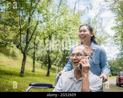 Fröhlicher, behinderter älterer Mann, der auf dem Smartphone spricht, während die Enkelin einen Rollstuhl im Park schiebt. Familienleben im Urlaub. Stockfoto