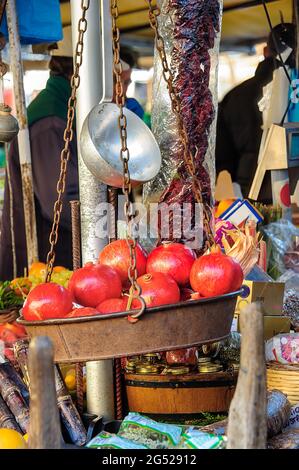 Granatäpfel in einer alten Metallwaage mit rostigen Ketten und trockenen Tomaten, Obst und Gemüse im Hintergrund. Stockfoto