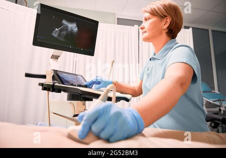 Sonografin, die Frau mit einem Ultraschallscanner untersucht. Der Arzt bewegt den Ultraschallaufnehmer am Abdomen des Patienten, während er auf das Display schaut. Konzept der medizinischen Untersuchung und Ultraschalldiagnostik. Stockfoto