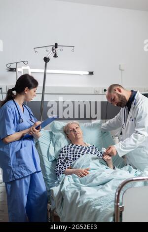 Die Krankenschwester notiert sich während der Konsultation einer älteren Frau im Klemmbrett, während der Arzt Daten vom Oxymeter über Puls, Blutpuls und Sauerstoffsättigung liest, die an einem kranken Patienten am Finger angebracht sind. Stockfoto