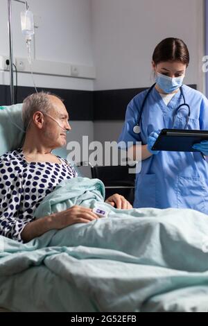 Medizinischer Assistent mit Schutzmaske, der die Behandlung des älteren Mannes beim Lesen von Notizen auf dem Tablet-pc überprüft. Kranke ältere Menschen, die im Bett liegen und mit der Krankenschwester über die Diagnosekompetenz mit angebacktem infusionsbeutel sprechen. Stockfoto