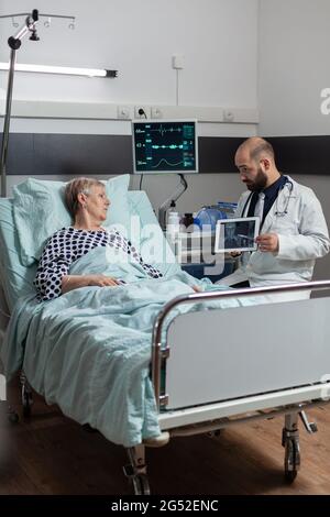 Der Arzt sitzt neben einem älteren Mann und erklärt die Diagnose eines Körpertraumas und zeigt Röntgenaufnahmen auf einem Tablet-pc im Krankenhauszimmer. Reifer Patient in der Klinik atmet es Hilfe aus Sauerstoffmaske. Stockfoto