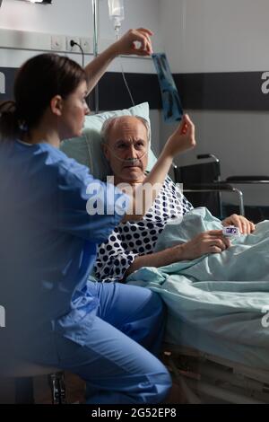 Alte Patientin mit Lungenerkrankung, die mit einer im Krankenhausbett liegenden Sauerstoffmaske atmet, lauschende Krankenschwester, die vor der Operation eine Röntgendiagnostik zeigt. Stockfoto