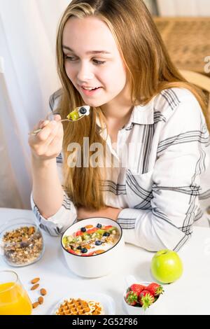 Junge lächelnde Frau, die zu Hause leckeres gesundes Müsli mit Obst und Joghurt zum Frühstück hat. Stockfoto