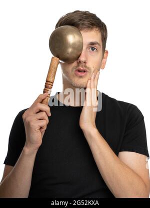 Junger, hübscher, großer, schlanker weißer Mann mit braunem Haar bedeckt sein Auge mit einer tibetischen Klangschale in schwarzem Hemd, isoliert auf weißem Hintergrund Stockfoto
