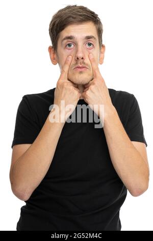 Junger, hübscher, großer, schlanker weißer Mann mit braunem Haar, das in einem schwarzen Hemd auf weißem Hintergrund auf seine Augen zeigt Stockfoto
